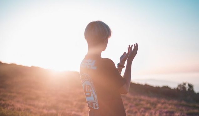 A person standing in a field at sunset, hands raised, with a soft glow illuminating the background.