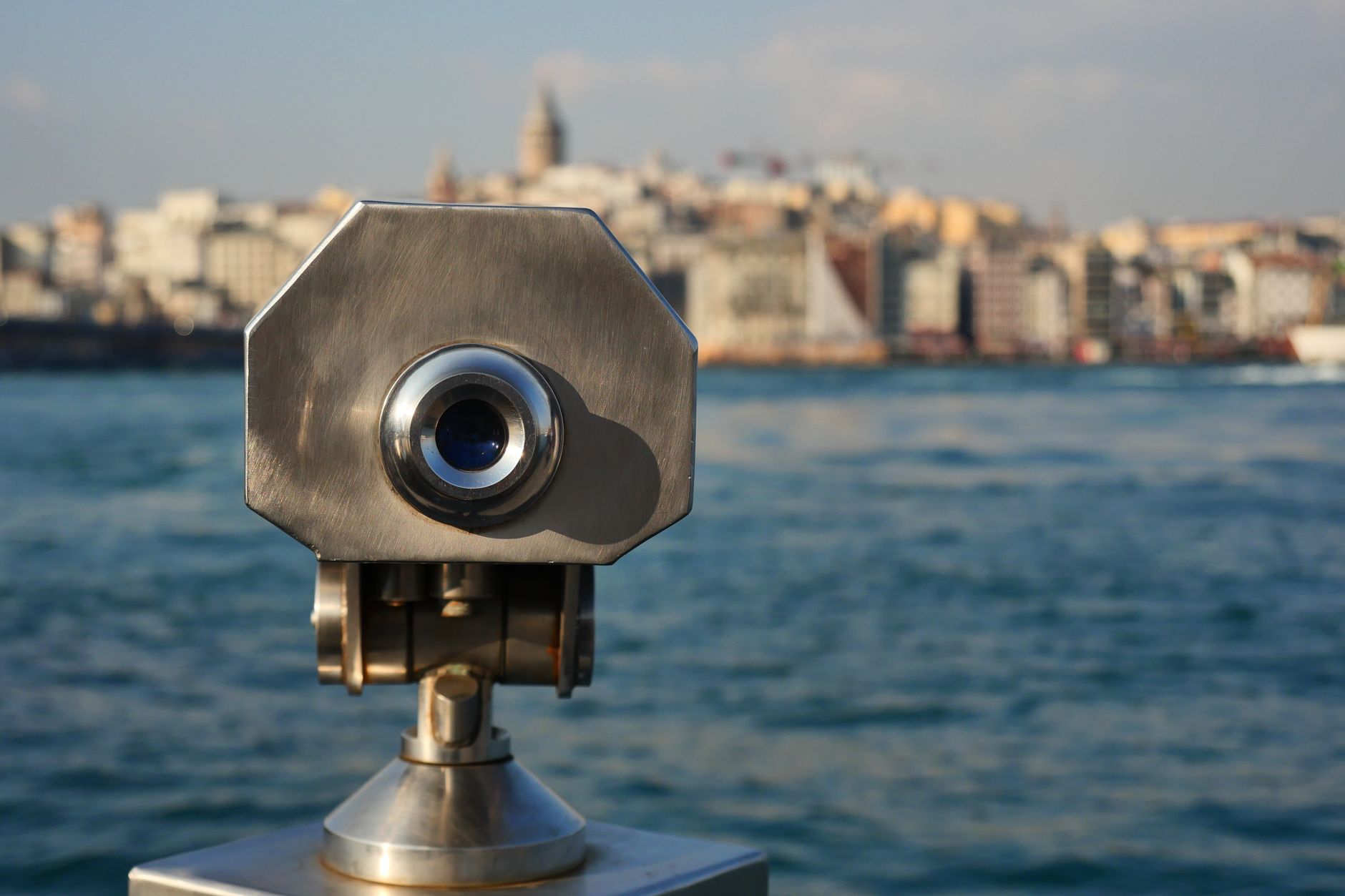 A metallic binocular viewer overlooking a waterfront with a blurry cityscape in the background.